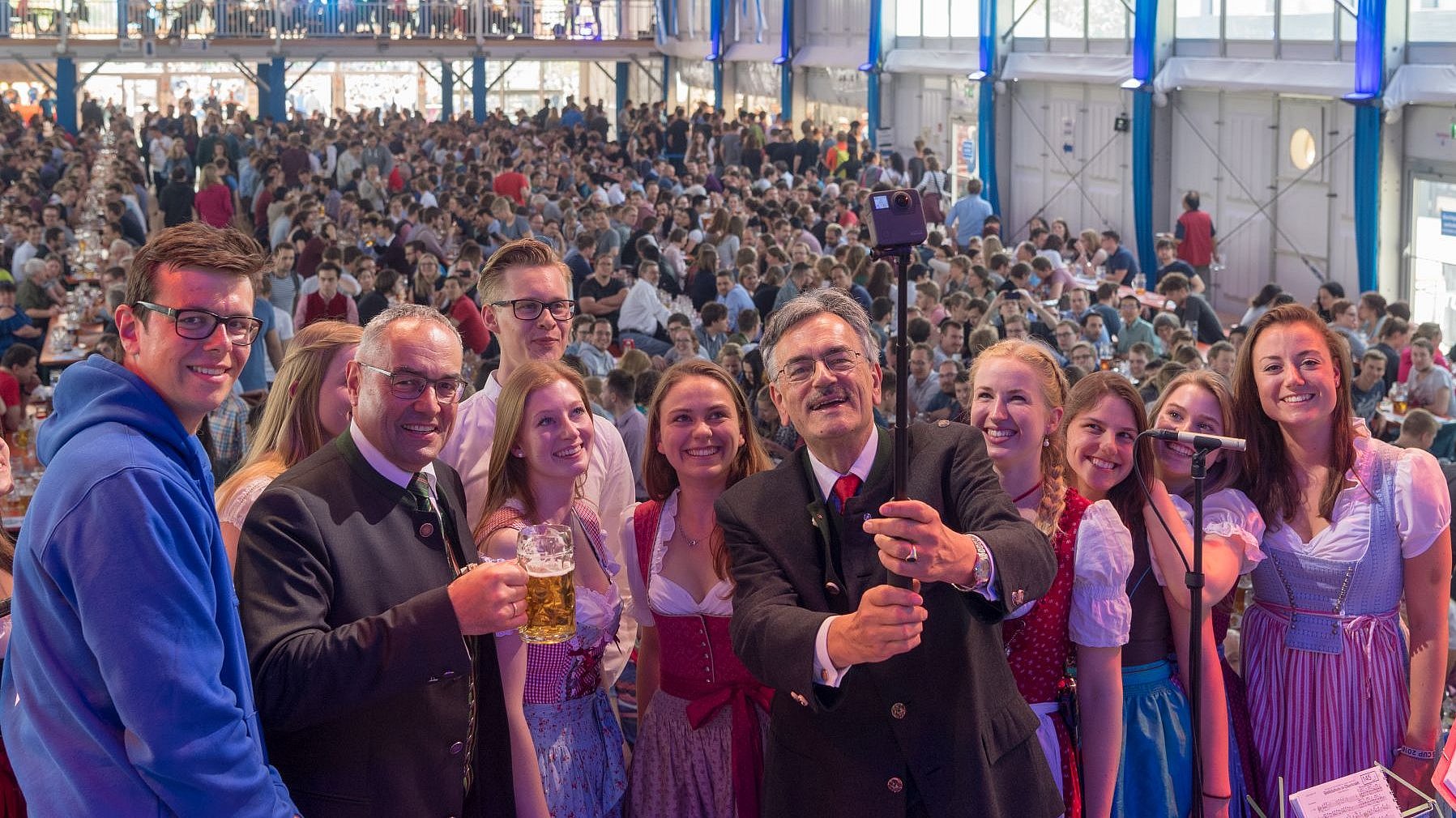 President Emeritus Wolfgang A. Herrmann, acting TUM President in the anniversary year 2018, and Chancellor Albert Berger (right) with students using a selfie stick in the marquee at the staff party in the anniversary year 2018, behind them numerous guests on beer benches. 
