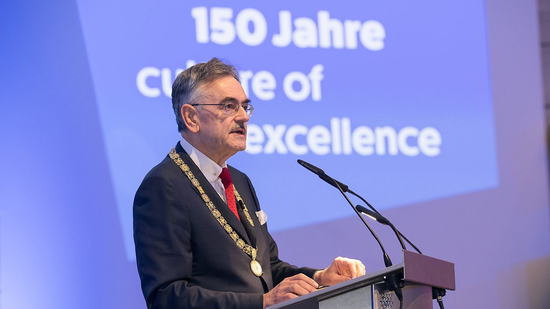  Wolfgang A. Herrmann speaks into a microphone. Behind him is a presentation with the words “150 years of culture of excellence.” 