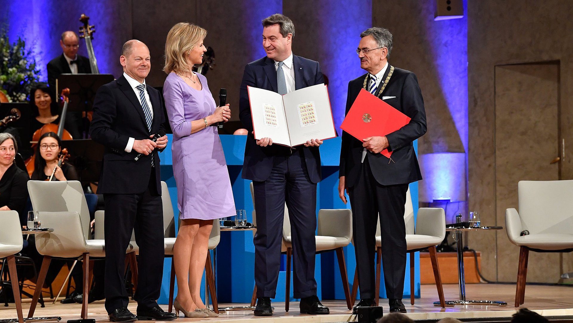 Olaf Scholz, Anouschka Horn, Markus Söder, and Wolfgang A. Herrmann (from left to right) stand together on a stage; Söder and Herrmann hold open folders in their hands. The occasion is the 2018 ceremony marking the 150th anniversary of the Technical University of Munich in the Hercules Hall of the Munich Residence.