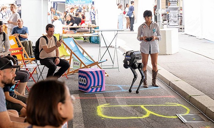 The image shows a knee-high humanoid robot and a young woman holding a remote control, standing in front of a seated audience.