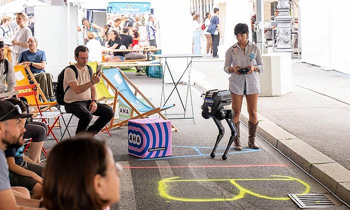 Das Bild zeigt einen kniehohen humanoiden Roboter und eine junge Frau mit Fernsteuerung in der Hand vor sitzendem Publikum
