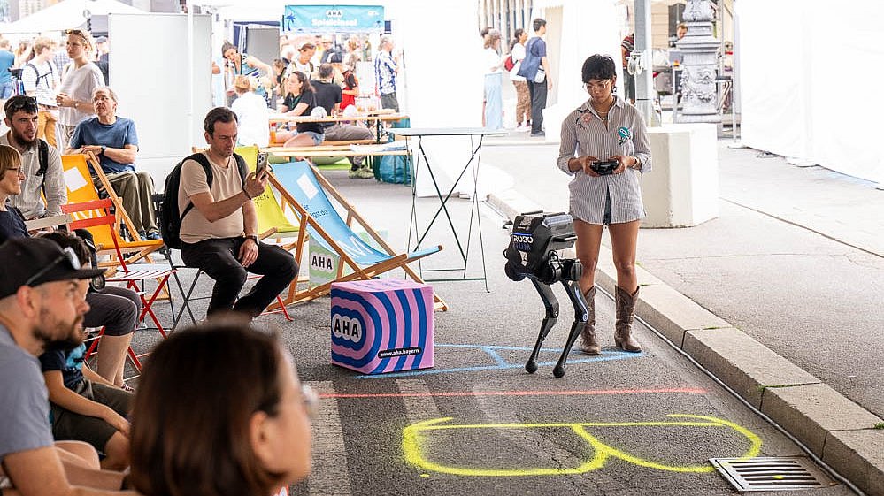 Das Bild zeigt einen kniehohen humanoiden Roboter und eine junge Frau mit Fernsteuerung in der Hand vor sitzendem Publikum