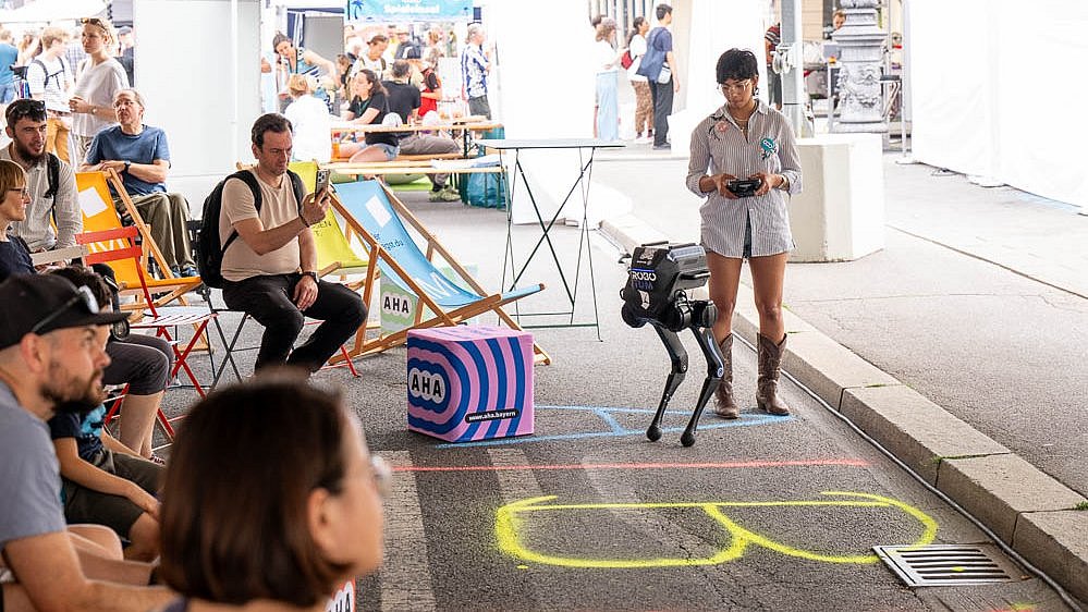 The image shows a knee-high humanoid robot and a young woman holding a remote control, standing in front of a seated audience.