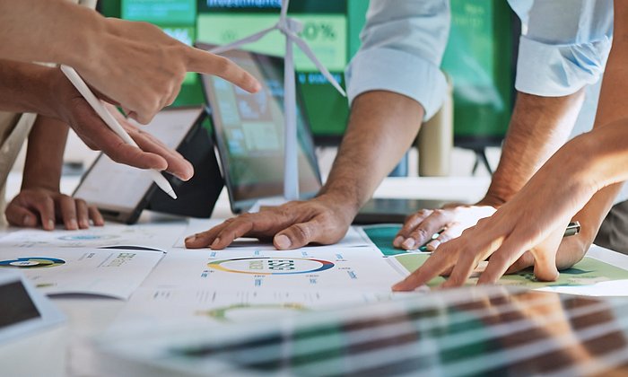 There are sheets of paper with various diagrams on a table. Several people are leaning on them, discussing the diagrams. However, only their hands are visible.