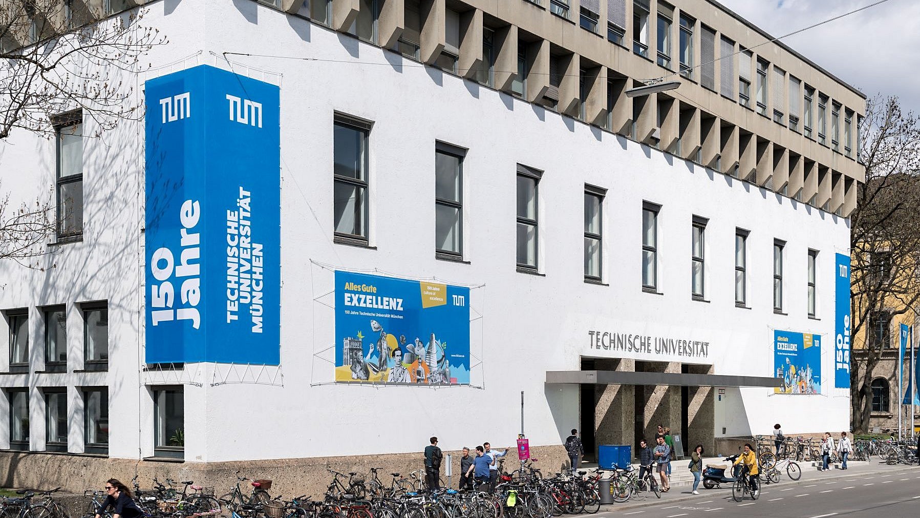 Exterior facade of the main campus of TUM in downtown Munich. Blue banners with white lettering commemorating the university's 150th anniversary in 2018 hang on the walls. 