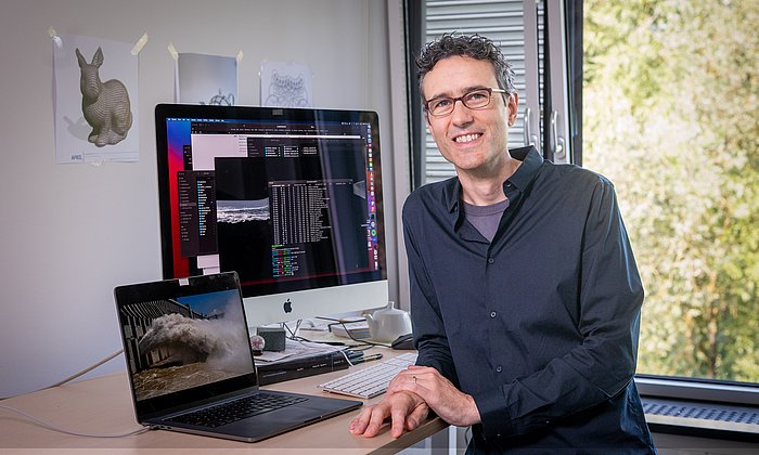 Prof. Nils Thuerey sits in front of two screens. The left screen shows a simulated wave motion. The right screen shows the corresponding source code.