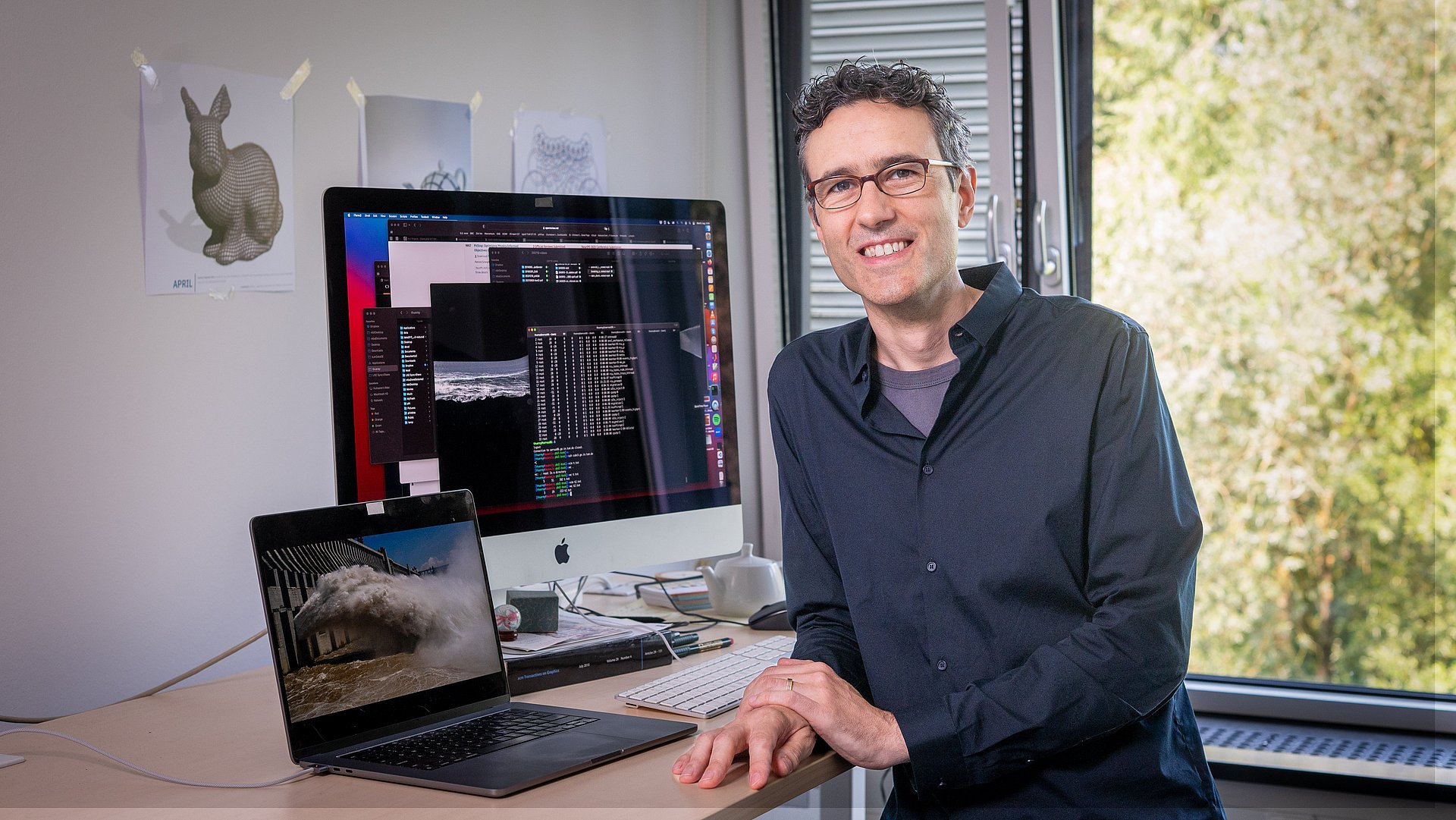 Prof. Nils Thuerey sits in front of two screens. The left screen shows a simulated wave motion. The right screen shows the corresponding source code.