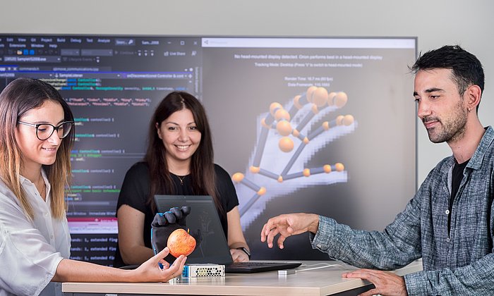 Three people standing around a table testing a prosthetic hand. The woman on the left is holding an apple, which the prosthetic hand is reaching for.