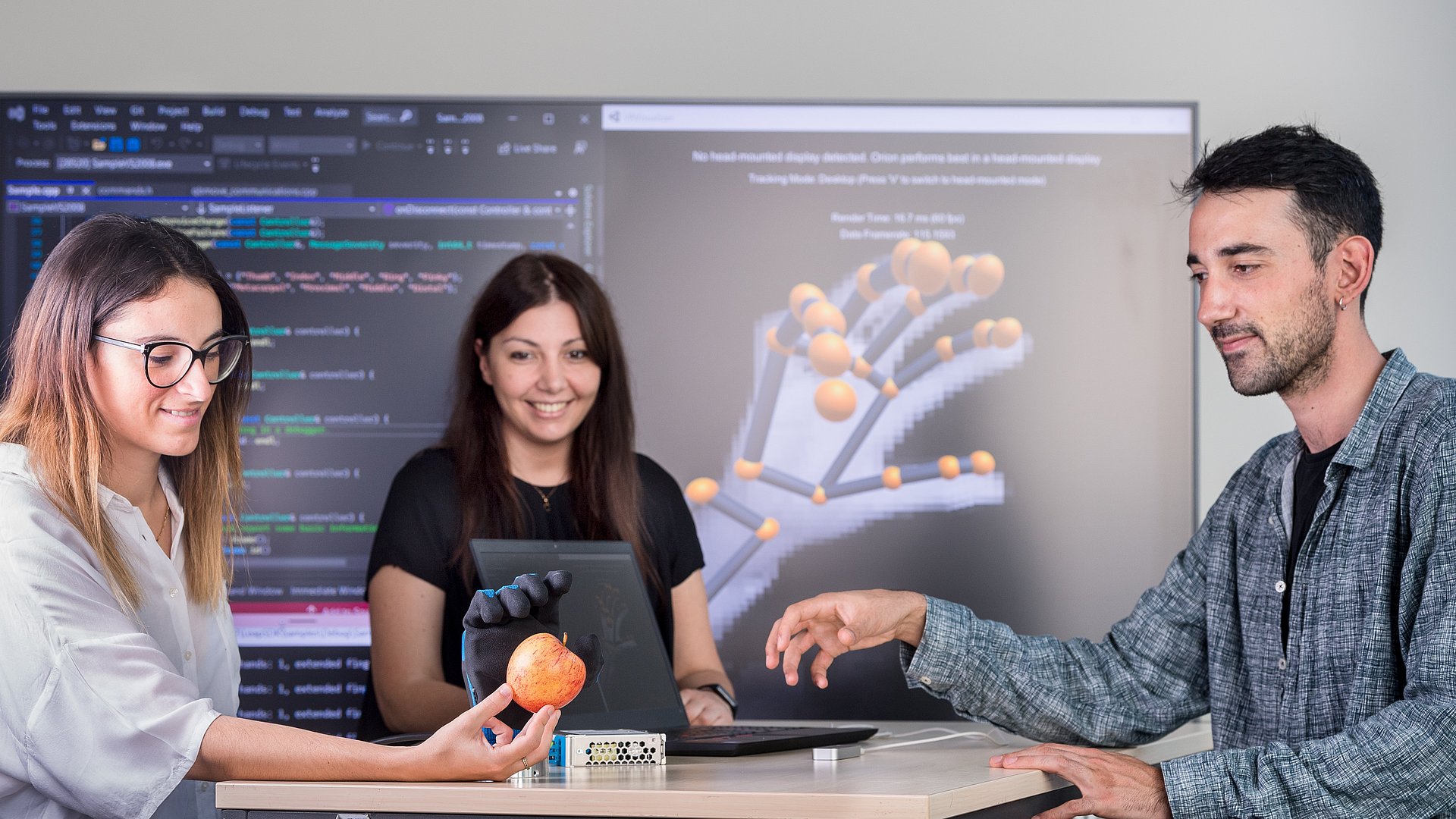 Three people standing around a table testing a prosthetic hand. The woman on the left is holding an apple, which the prosthetic hand is reaching for.
