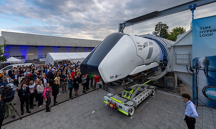 A Hyperloop Pod is on a transport rack in front of a concrete tube. Many guests are standing next to it.