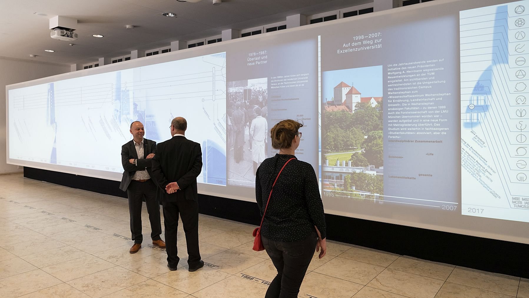  Installation of the exhibition “Zeitlupe” (Slow Motion) in the TUM matriculation hall with one female visitor and two male visitors.