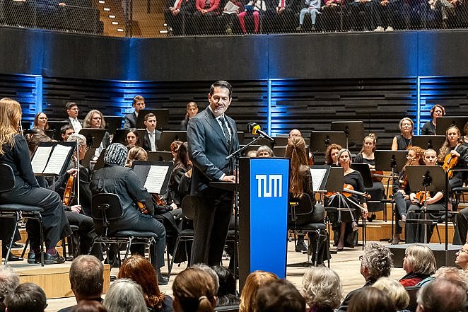 Auf der Bühne steht TUM-Präsident Prof. Hofmann und hält eine Begrüßungsrede. Hinter ihm die Musiker des SEM-Orchesters