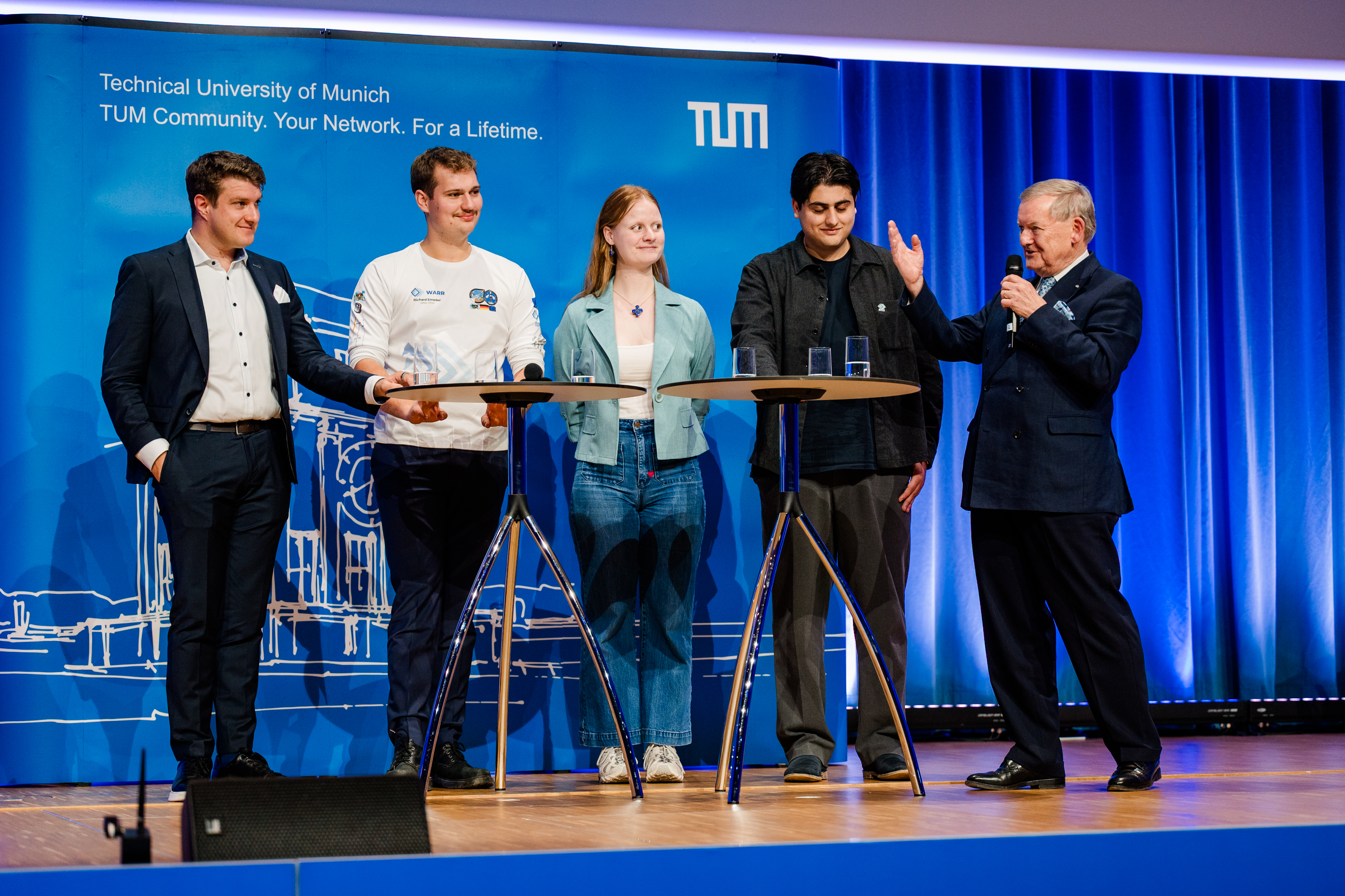 Five people standing on a stage in front of a blue background at two bar tables, from left to right: Josef Fleischmann, Richard Emeder, Mareike Ufer, Ceyhun Aydin, and Robert Schmucker; Mr. Schmucker is speaking into a handheld microphone, facing the other four, and gesturing with his right hand.